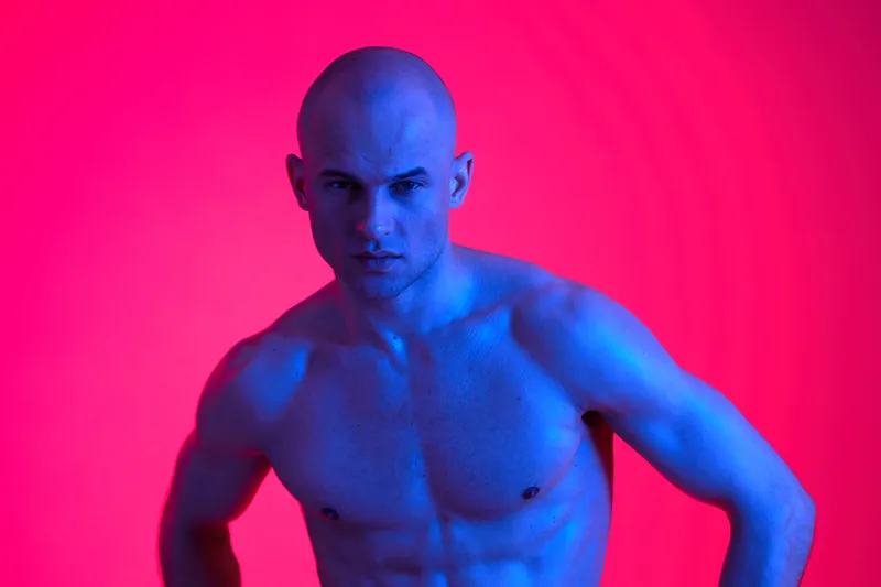 Muscular man posing confidently under colorful lights, showcasing his strength and determination. The vibrant background adds a dynamic atmosphere to the image.
