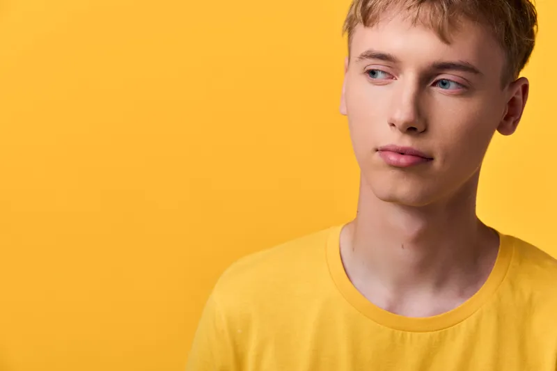 Young man wearing a yellow shirt stands against a bold yellow backdrop, conveying contemplation and calm in a minimalist studio scene with strong color and balanced lighting for a modern, thoughtful