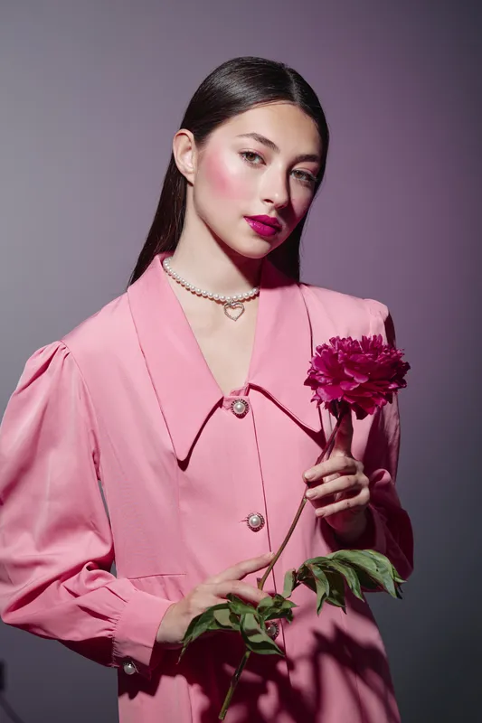 Young woman in pink vintage coat poses with a bloom, styled for beauty fashion creative portrait on gradient purple. Concept for elegance, softness, and glamour.