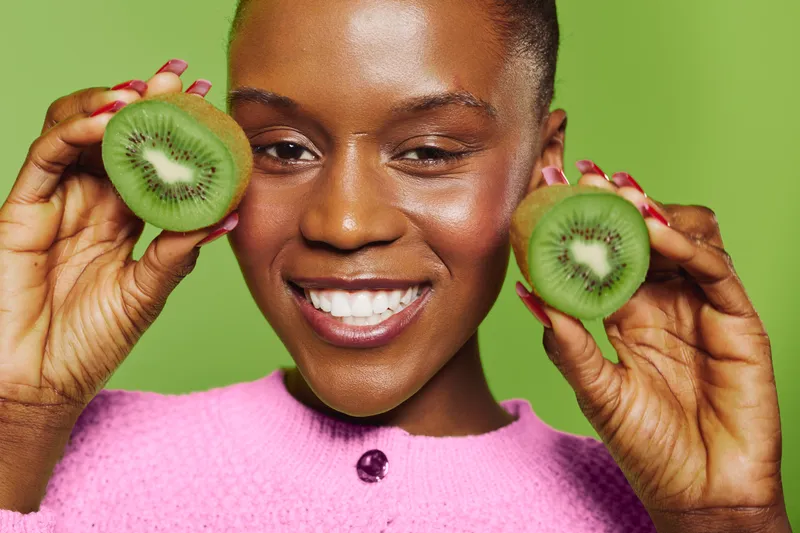 Smiling black woman with dark skin holds two halves of fresh kiwi fruit near her face against green background. Concept of healthy lifestyle, nutrition, and natural beauty.