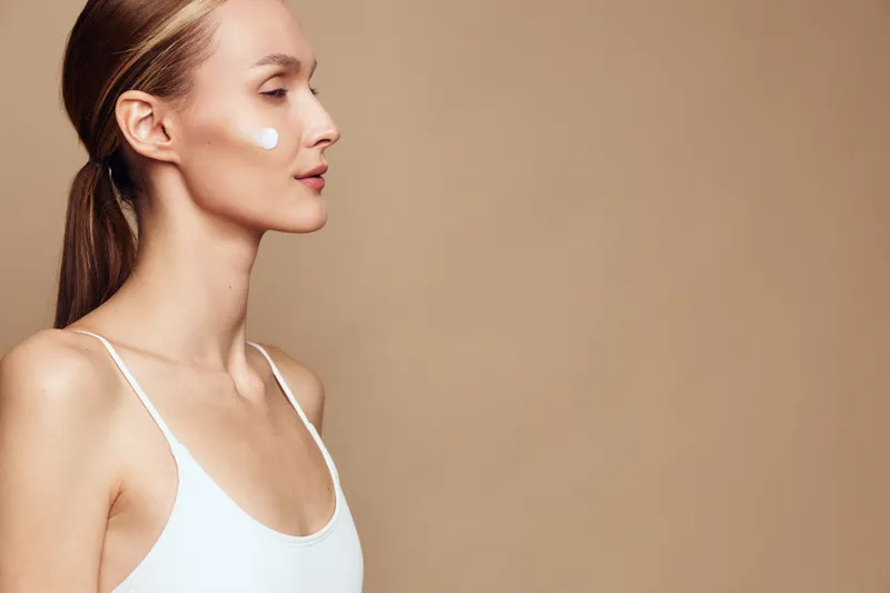 Young woman with natural skin applies moisturizing cream on cheek in white tank top isolated on beige background with closed eyes and relaxed expression for skincare concept.
