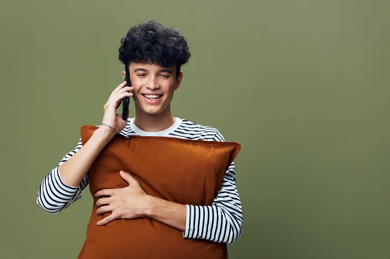 smiling man talking on phone holding brown pillow, dressed in striped shirt with curly hair, isolated on green background, casual lifestyle moment