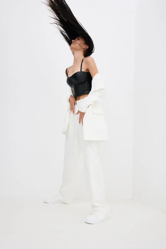 Young woman with long black hair flipping her hair, dressed in stylish black corset and white pants, posing confidently in bright minimalist studio with white background.
