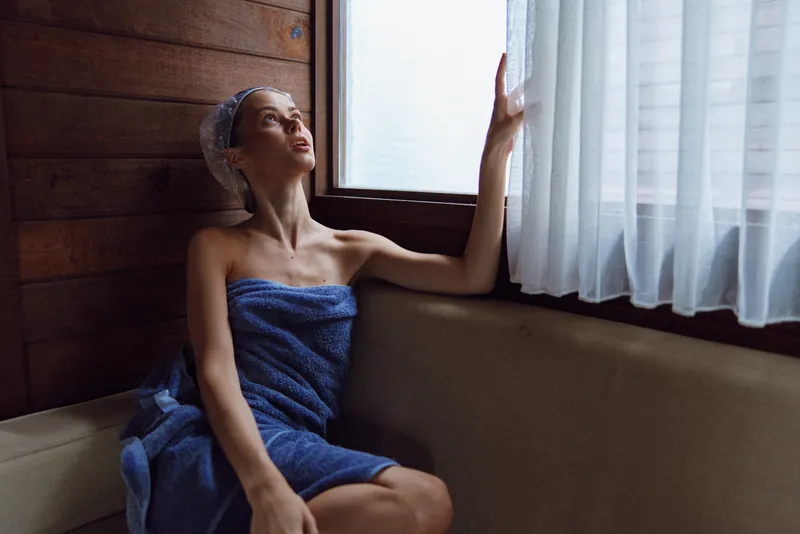Young woman wrapped in a blue towel sits by a window in a wooden sauna, relaxing and enjoying natural light with a contemplative expression and shower cap.