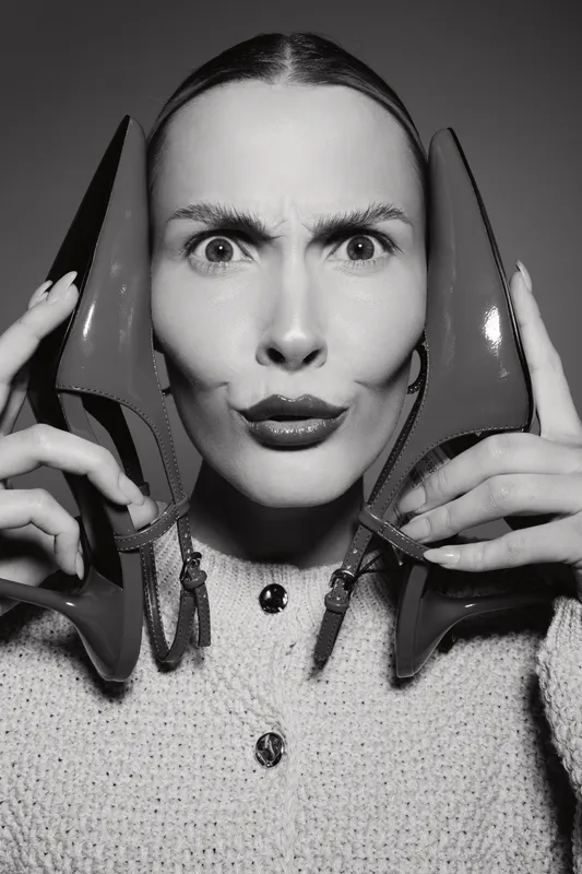 Young white woman with pale skin holds red high heels near face with playful expression in studio black and white portrait concept for fashion.