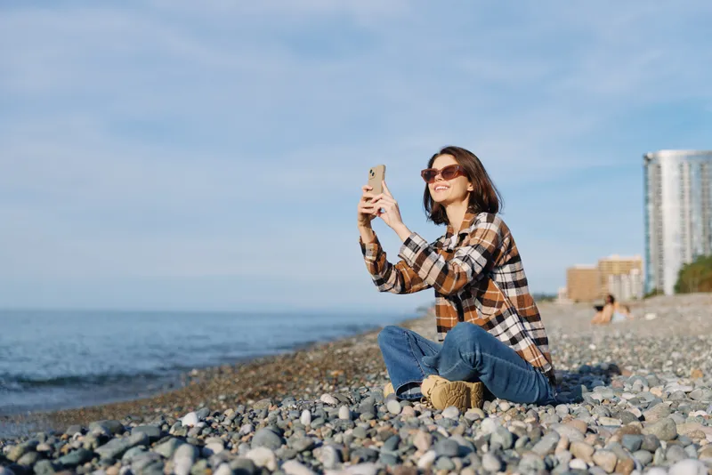 Woman taking a selfie on a rocky beach, wearing a plaid shirt and jeans, sunglasses on, close to the water with distant buildings, a casual outdoors moment for lifestyle and travel