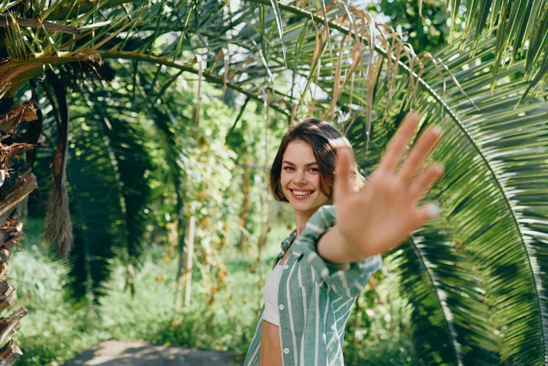 Smiling woman in a tropical garden waves at the camera, capturing a candid, sunny moment among lush palm leaves and vibrant greenery.