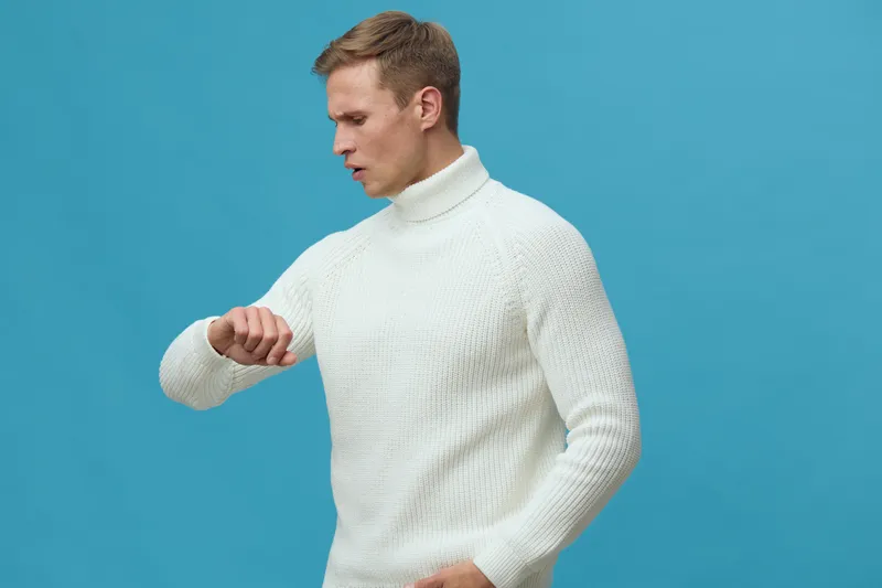 Man wearing white turtleneck sweater looks at watch isolated on solid blue background studio portrait. People fashion casual style winter clothing concept
