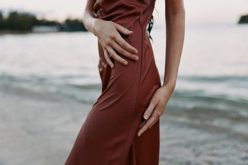 Close up of a person in a warm brown dress by a calm shoreline, soft light highlights the fabric drape and silhouette, creating a serene, elegant mood and subtle detail.