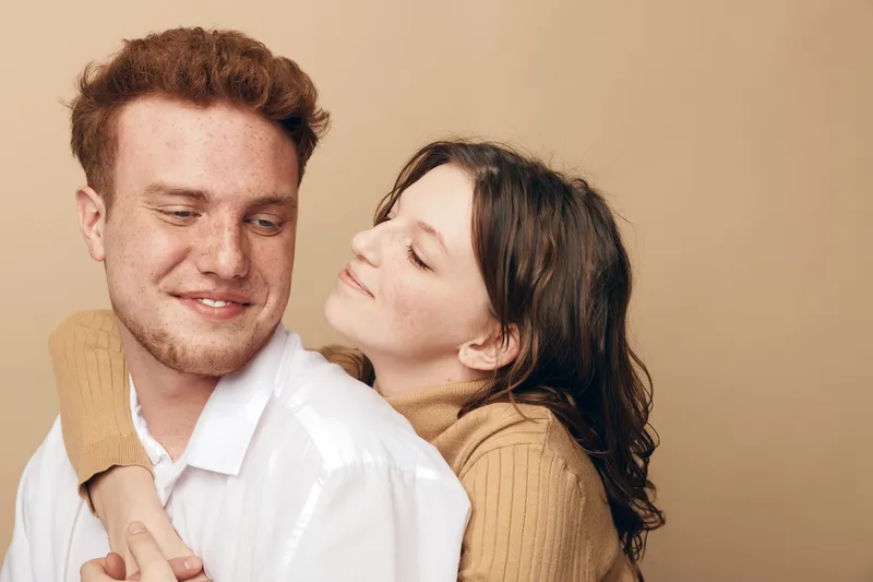 Young couple in casual clothes embraces tenderly against beige background. Woman hugs man from behind with closed eyes. Concept of love, affection, intimacy, and relationship.