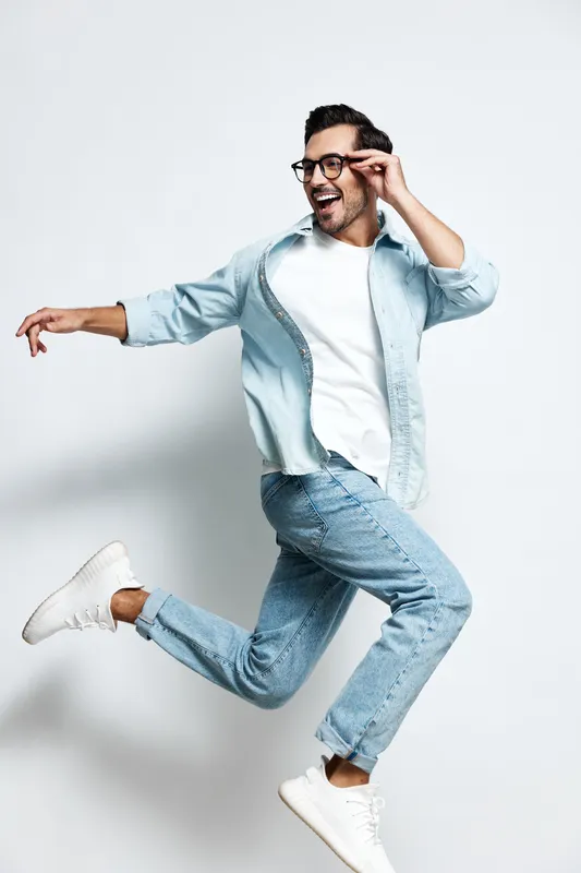 Full body joyful young man wearing glasses and a light blue shirt, jumping in excitement while looking at the camera against a clean gray background. People lifestyle concept
