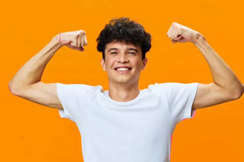 young man flexing muscles confidently against bright orange background, smiling with casual white t-shirt, showing strength and positive energy