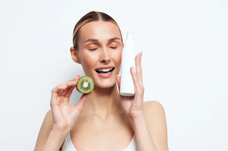 A young woman with clear skin holds a kiwi slice and a white cosmetic bottle, smiling with closed eyes. Concept for natural skincare, beauty, and healthy lifestyle products.