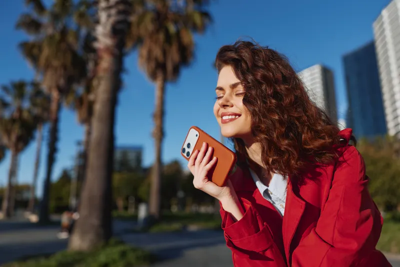 Smiling woman in a red coat enjoys a sunny outdoor moment, holding a smartphone near her ear, with palm trees and modern buildings in the background with urban energy