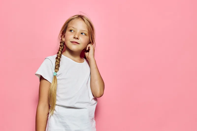 Young girl with long blonde hair in braids wears a white t-shirt and looks thoughtfully to the side on a pink background. Studio portrait for childhood concept.