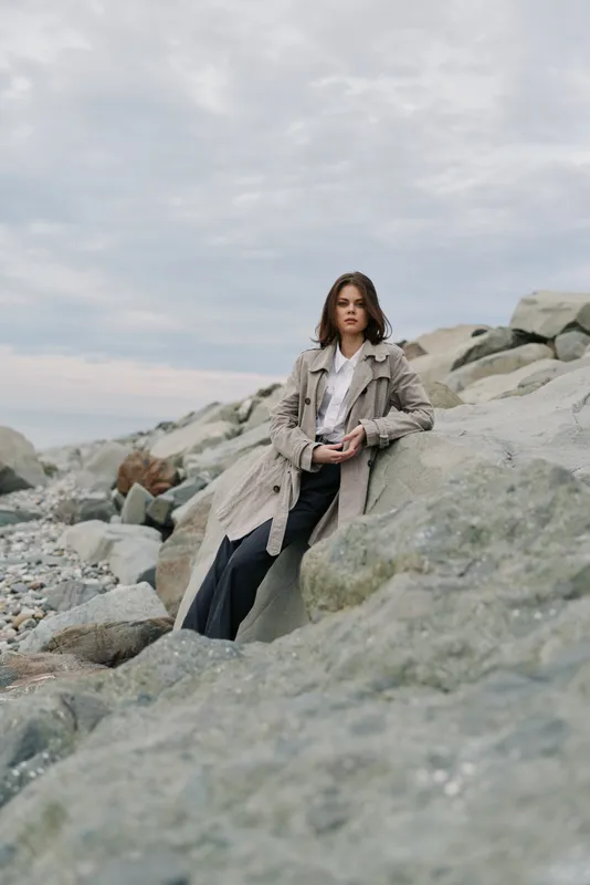 Young woman in casual coat sits on rocky shore under cloudy sky. Calm natural environment creates mood of relaxation and peaceful solitude near the sea.
