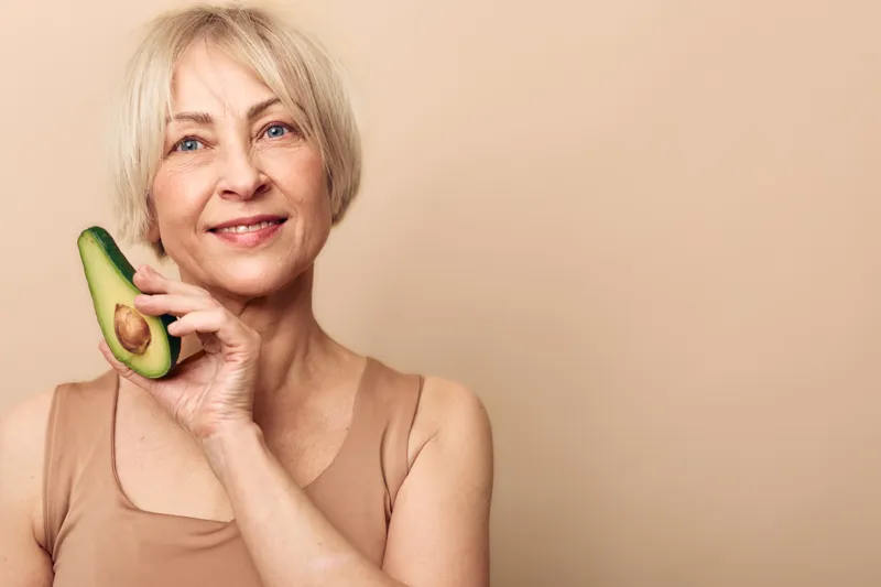 Smiling mature woman with short blonde hair holding half of ripe avocado near face on beige background. Concept of healthy diet, natural skincare, wellness, and positive aging.
