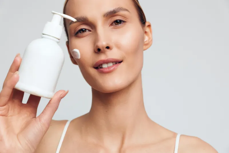 Young woman holding white lotion bottle applying cream on face closeup skincare routine natural skin beauty treatment clean background wellness concept