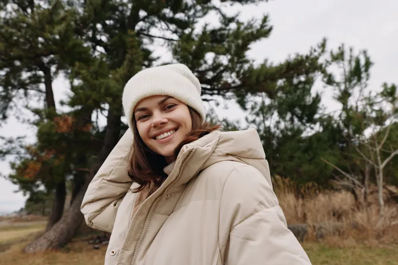 Smiling woman in a warm beige coat and white beanie posing outdoors with trees and grass in the background, capturing casual winter style.