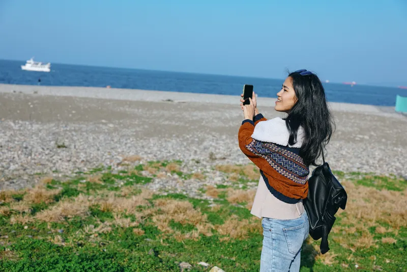 Beach scene with a woman on a sandy coastline capturing a moment with a smartphone, casual attire, backpack, and ocean view under a clear blue sky for stock