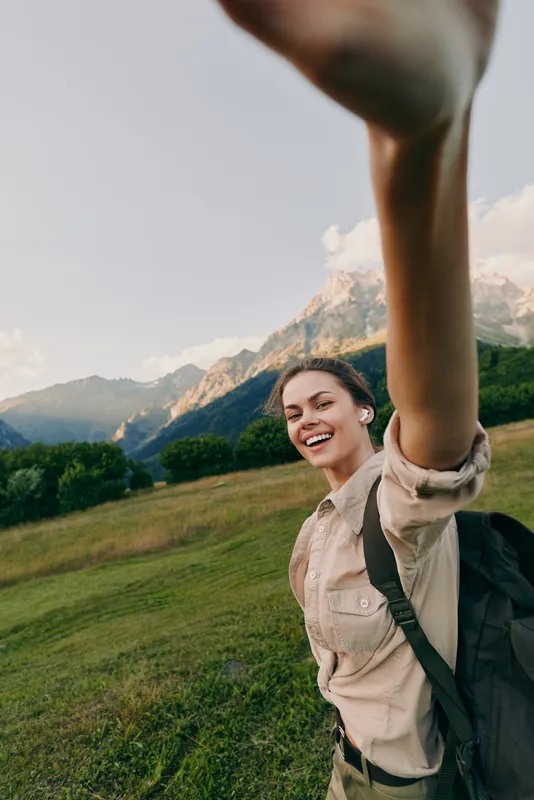 Young woman with a backpack smiles joyfully while taking a selfie in a mountainous meadow under a clear sky during daytime.