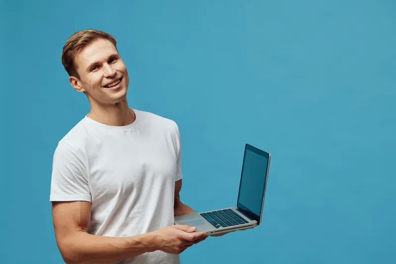 Smiling young man holding open laptop computer in casual white t-shirt against blue background, showcasing technology use, modern lifestyle, and positive emotions. Studio portrait, people concept, cas