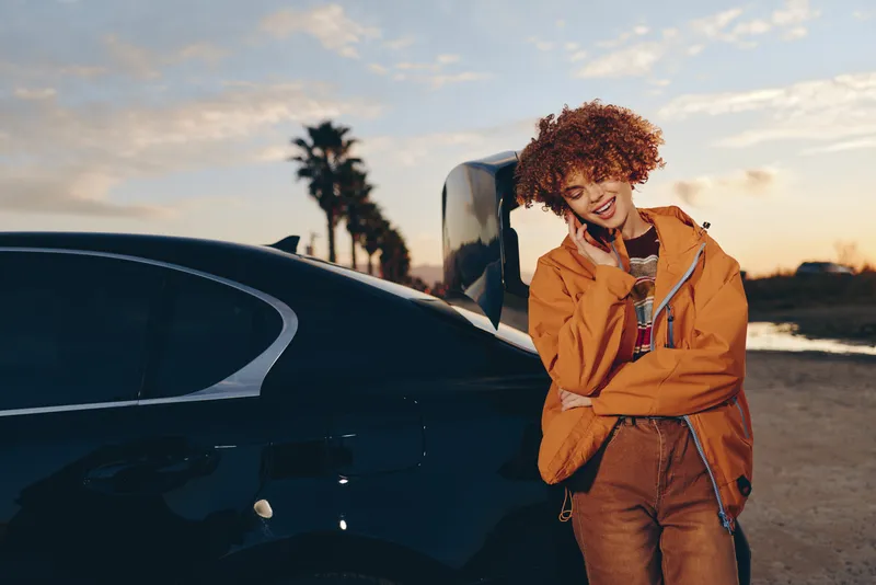 Woman lifestyle outdoors at sunset, standing by car near beach road, smiling, curly hair, casual orange jacket, relaxed arms crossed, warm golden light, soft sky, evening breeze