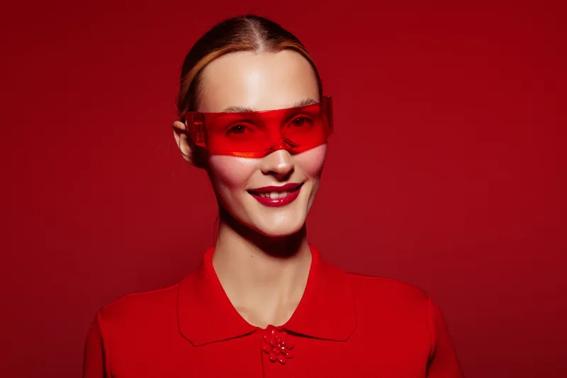 Smiling woman with red sunglasses and red outfit poses against red background. Studio portrait with bright lighting and fashion concept focused on color coordination and style.