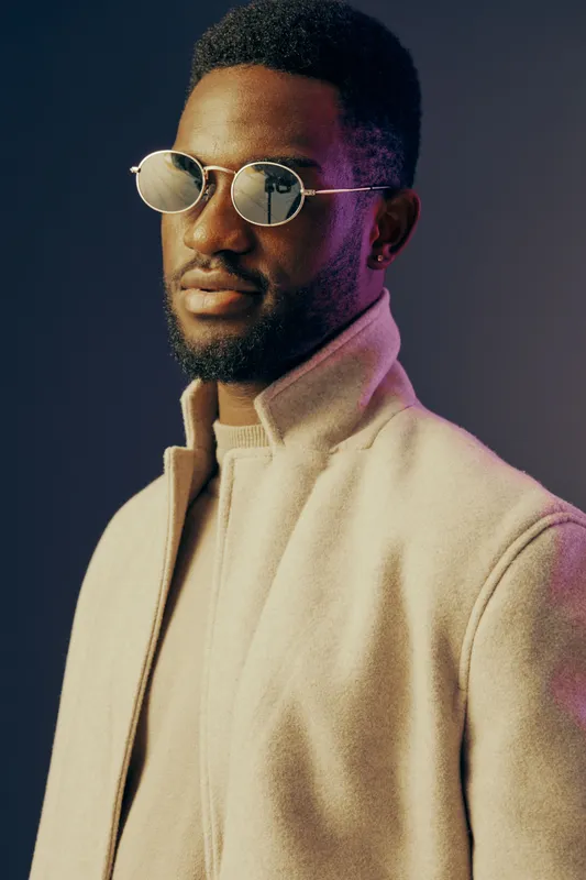 Stylish black male model wearing sunglasses and a modern beige coat, exuding confidence against a colorful gradient background