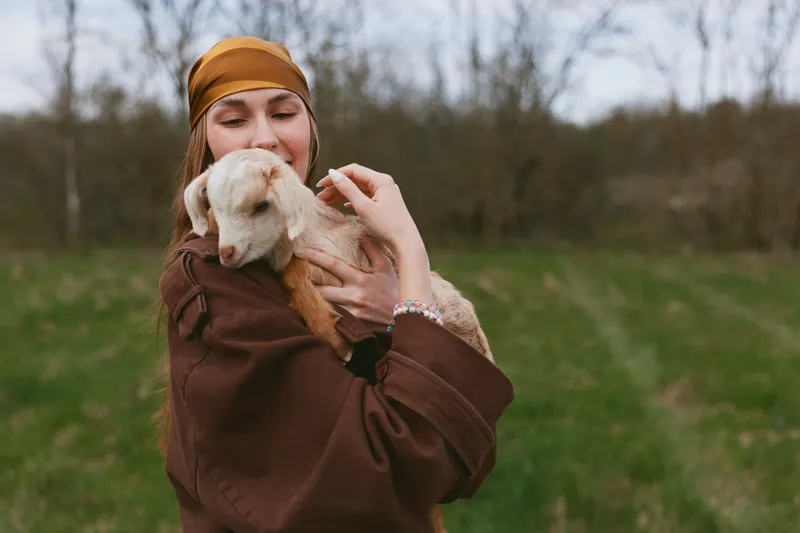 Young woman wearing a brown jacket and headscarf gently holds a baby goat in a grassy field during a calm outdoor lifestyle moment in early spring.