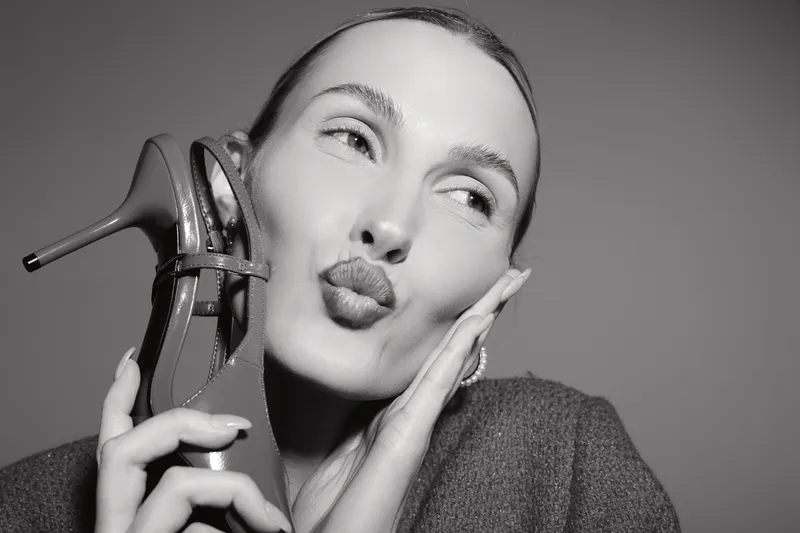 Young white woman with fair skin puckers lips holding high heel shoe near face in studio. Concept of beauty, fashion, and playful mood.