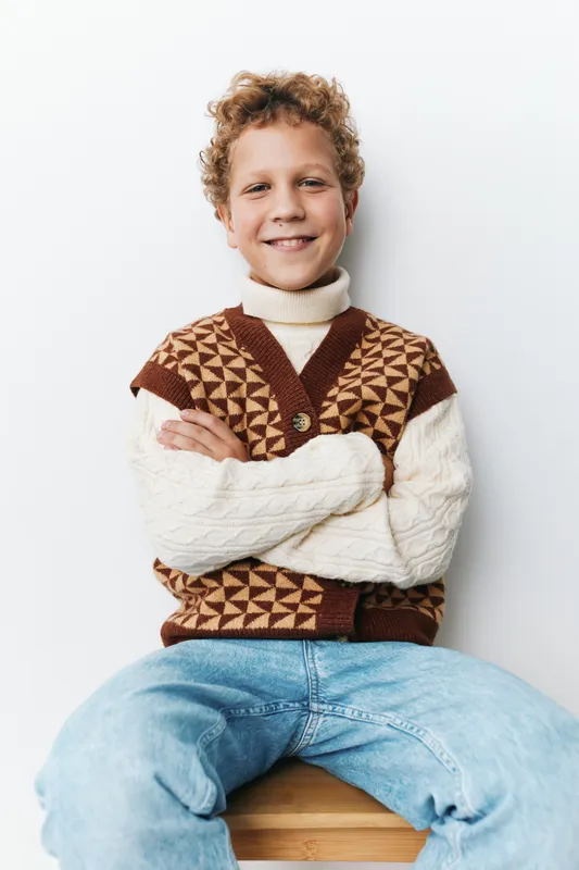 Portrait of a cheerful boy seated against a white wall, wearing a geometric knitted vest over a cream turtleneck and blue jeans, exuding friendly charm.