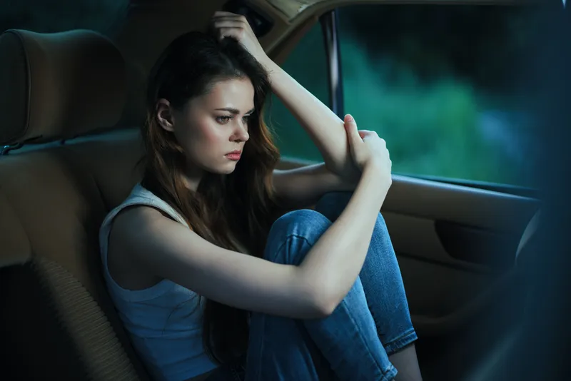 Young woman sitting alone in a car, expressing sadness, wearing casual clothes, with a moody green background, reflecting feelings of isolation and contemplation.