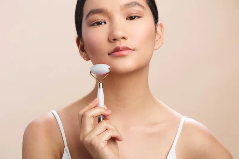 Asian woman uses a facial roller in a calm studio setting. She wears a light top and looks relaxed, presenting a serene beauty routine moment with soft lighting.