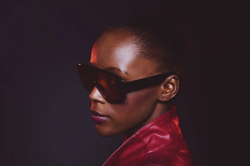 Profile of a confident dark skinned African woman wearing large stylish sunglasses and a red leather jacket against a dark background. Concept of fashion, elegance, and modern style.