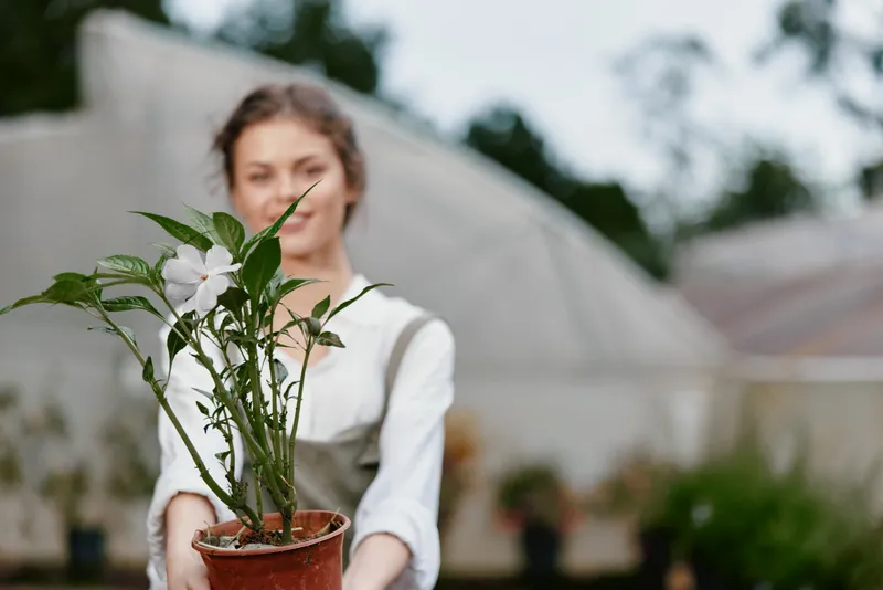 Young woman joyfully presenting a potted flower in a greenhouse, emphasizing connection with nature, gardening passion, and vibrant greenery