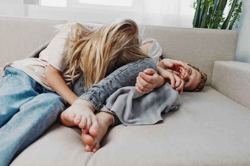 Two children lying on a sofa playfully interacting, with one child covering the other's face. Casual home setting with natural light and relaxed atmosphere, concept of childhood fun and sibling bond.