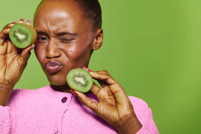 Close-up of dark skinned African woman in pink sweater holding two halves of kiwi fruit near face with playful expression on green background. Concept of healthy eating and natural lifestyle.