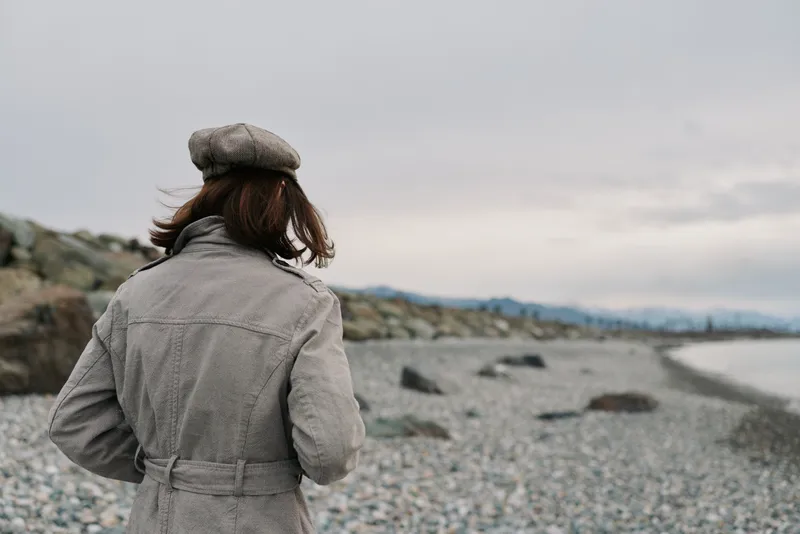 Woman in coat and beret stands on rocky beach looking at cloudy sky and distant mountains. Concept of solitude, travel, autumn fashion, and nature landscape by the sea.