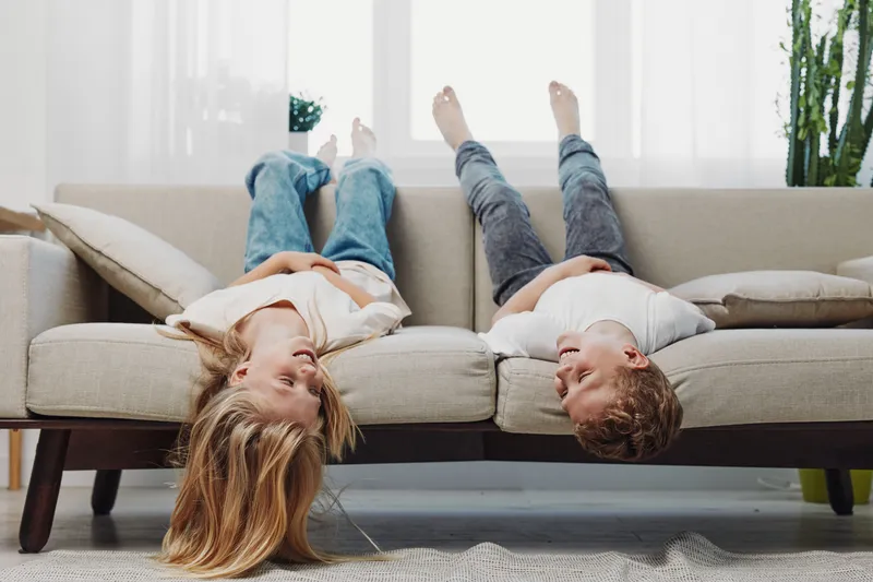 Two children lying upside down on a beige sofa in casual clothes, smiling and playing in a bright living room with natural light and cozy cushions around.