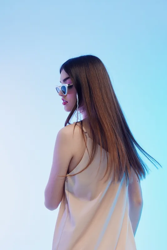 Young woman with long hair wearing sunglasses and a sleeveless top poses sideways on a blue background. Concept of woman beauty fashion and modern style.