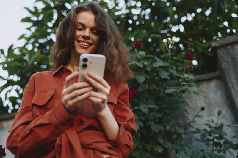 Young woman in a rust shirt smiles at her phone while standing in a garden with blooming roses, capturing a candid moment of everyday connection and joy