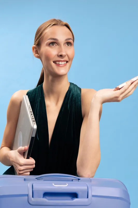 Smiling woman holding laptop and passport stands behind blue suitcase on light blue background. Concept for travel, vacation, business trip, and holiday planning with positive mood.