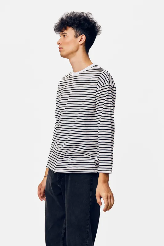 Young man wearing a striped long-sleeve shirt and dark pants standing sideways against a light background, expressing calm and thoughtful mood in casual attire.