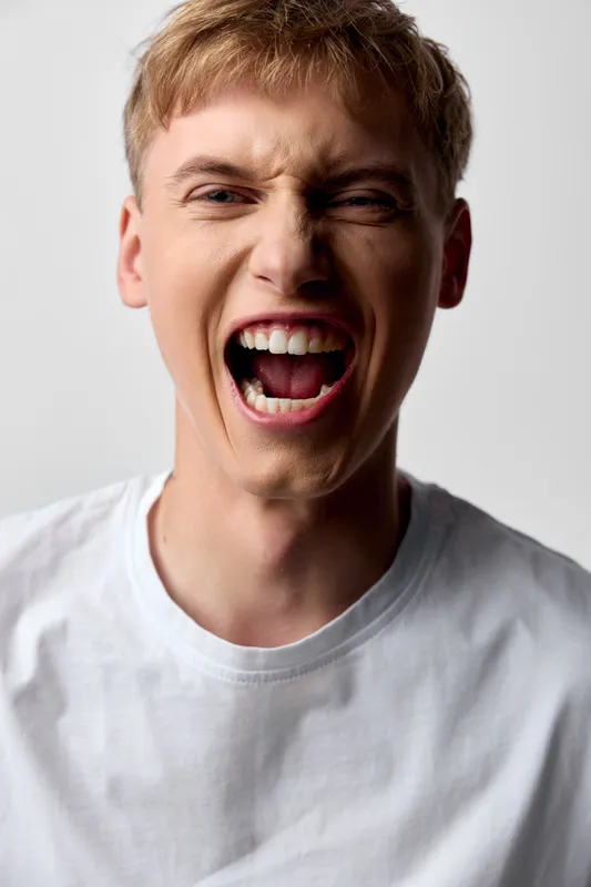 Close up of a man with short blond hair, mouth wide open in a powerful shout, wearing a white tee, studio lighting and bold contrast capturing raw energy and intense emotion