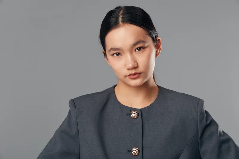 Portrait of confident Asian woman with natural makeup and dark hair tied back wearing a gray buttoned blouse standing against plain gray background in studio lighting.
