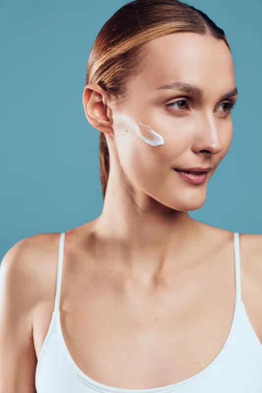 Young woman with clean skin applying face cream on cheek, natural beauty, smooth complexion, skincare routine, minimal makeup, blue background, healthy lifestyle concept