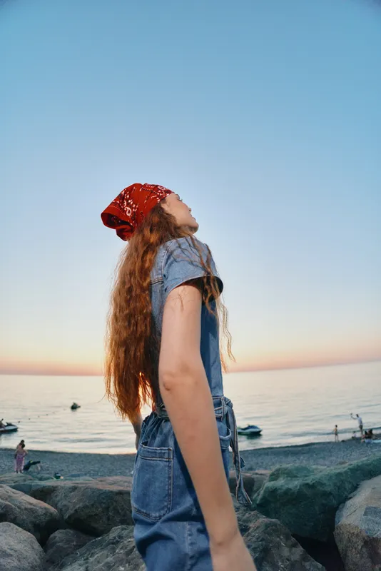 Lifestyle woman in modern boho-western denim street style with a red bandana, long hair flowing, enjoying the coastal sunset in warm film color tones.