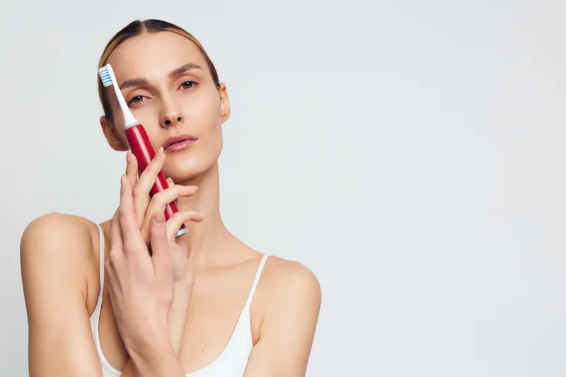 A young woman with clear skin holds a red electric toothbrush near her face against a plain light background. Concept for dental care, hygiene, beauty, and personal routine.