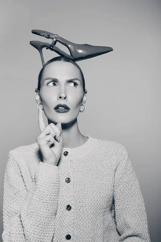 A caucasian woman with pale skin and dark lipstick balances a high heel shoe on her head while thinking. Concept of fashion, creativity, and style.